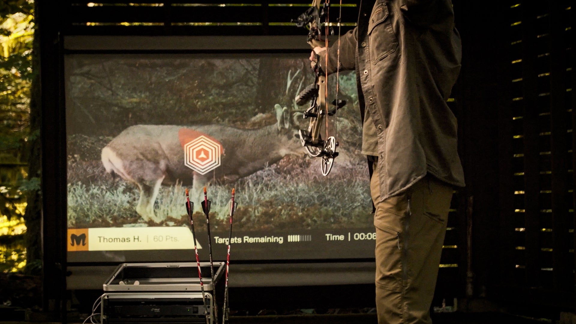 Person holding a bow and arrow in front of a large screen displaying a deer and minum archery simulator.